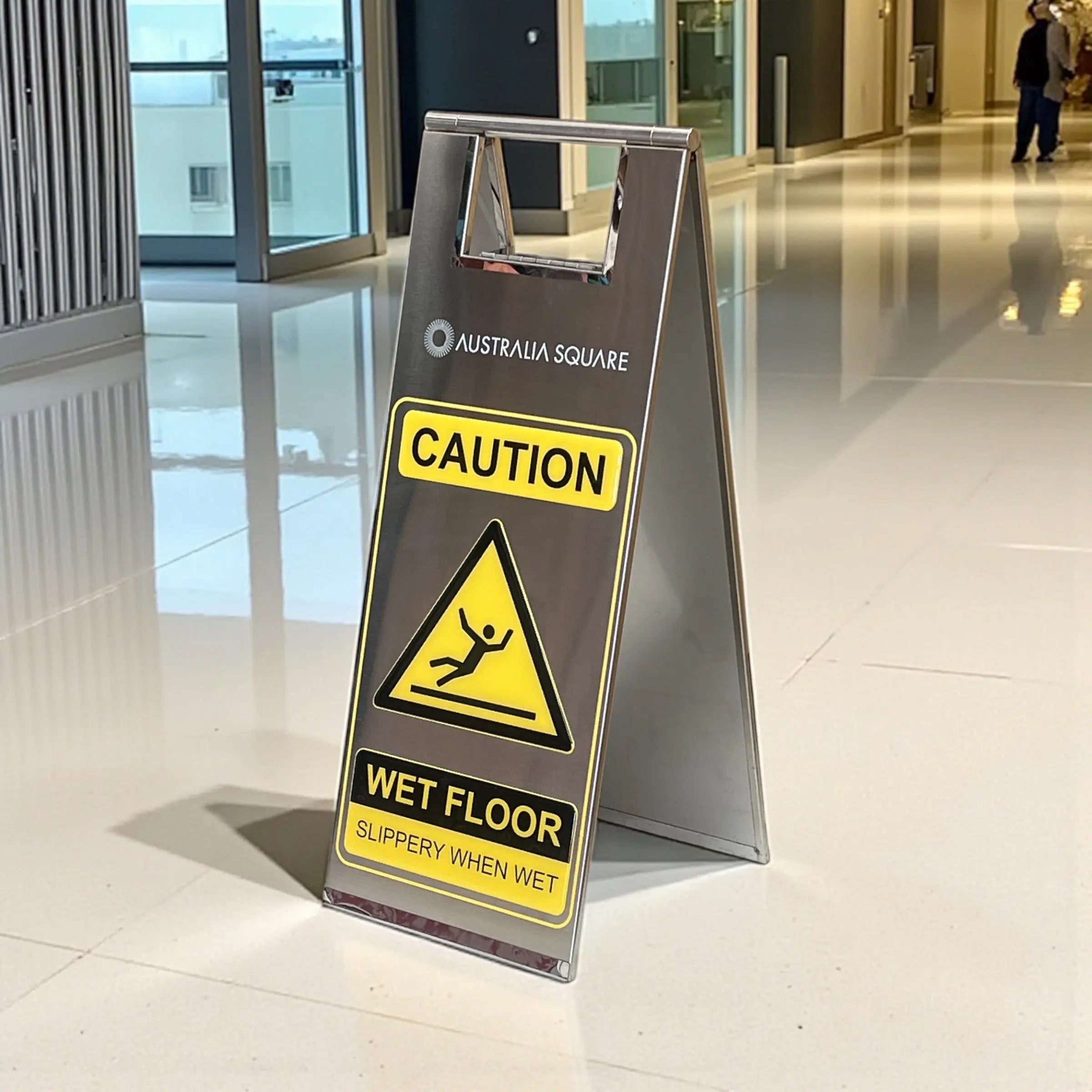 Stainless steel wet floor sign in silver, folding A-frame caution sign with yellow WET FLOOR panel, shown in a commercial building lobby